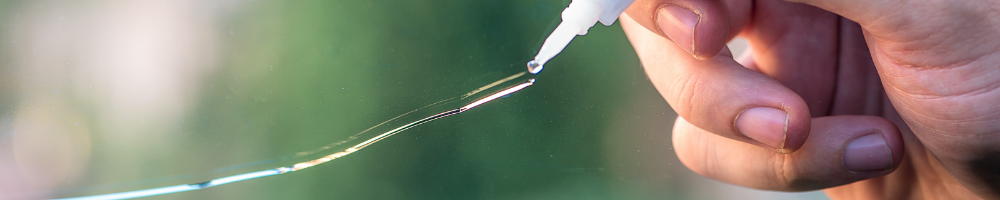 A close-up image of a person applying repair resin to a crack in a car windshield using a dropper, with a green, blurred background.