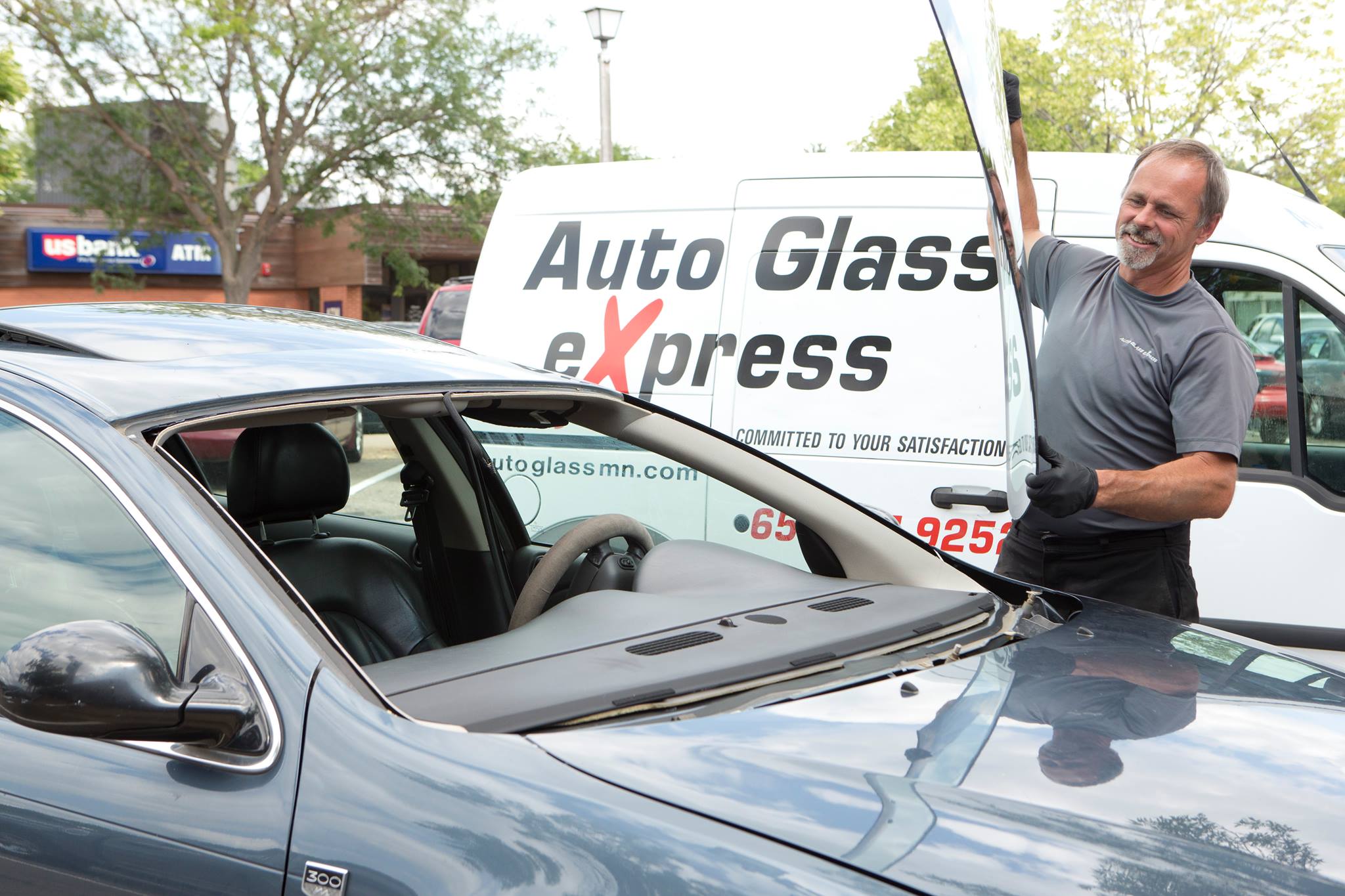 A smiling technician from Auto Glass eXpress as he replaces a car windshield. He is holding a large sheet of glass in his gloved hands, positioning it to fit into the frame of a dark-colored vehicle. The technician is wearing a grey shirt and black gloves, working efficiently and carefully. In the background, a white company van with the Auto Glass eXpress logo and contact information is parked, indicating the mobile service. The scene takes place outdoors, with trees and buildings visible in the background, highlighting the professional and convenient service provided by Auto Glass eXpress.
