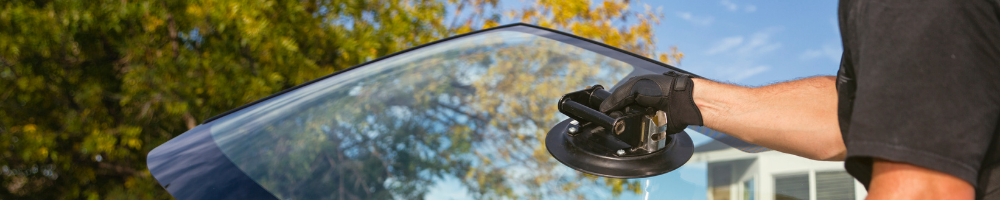 A person wearing black gloves using a suction cup tool to lift and remove a car windshield, with green trees and a blue sky in the background.