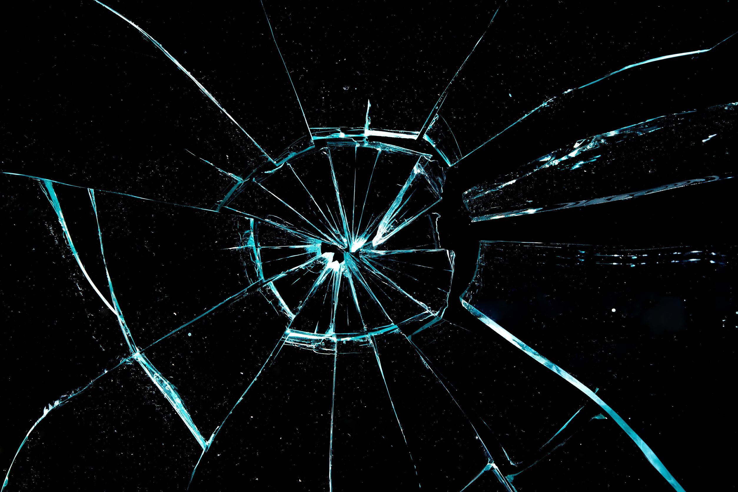 Close-up image of a shattered piece of glass with a spiderweb pattern of cracks radiating from the center, set against a dark background. | Can you drive with a cracked windshield?