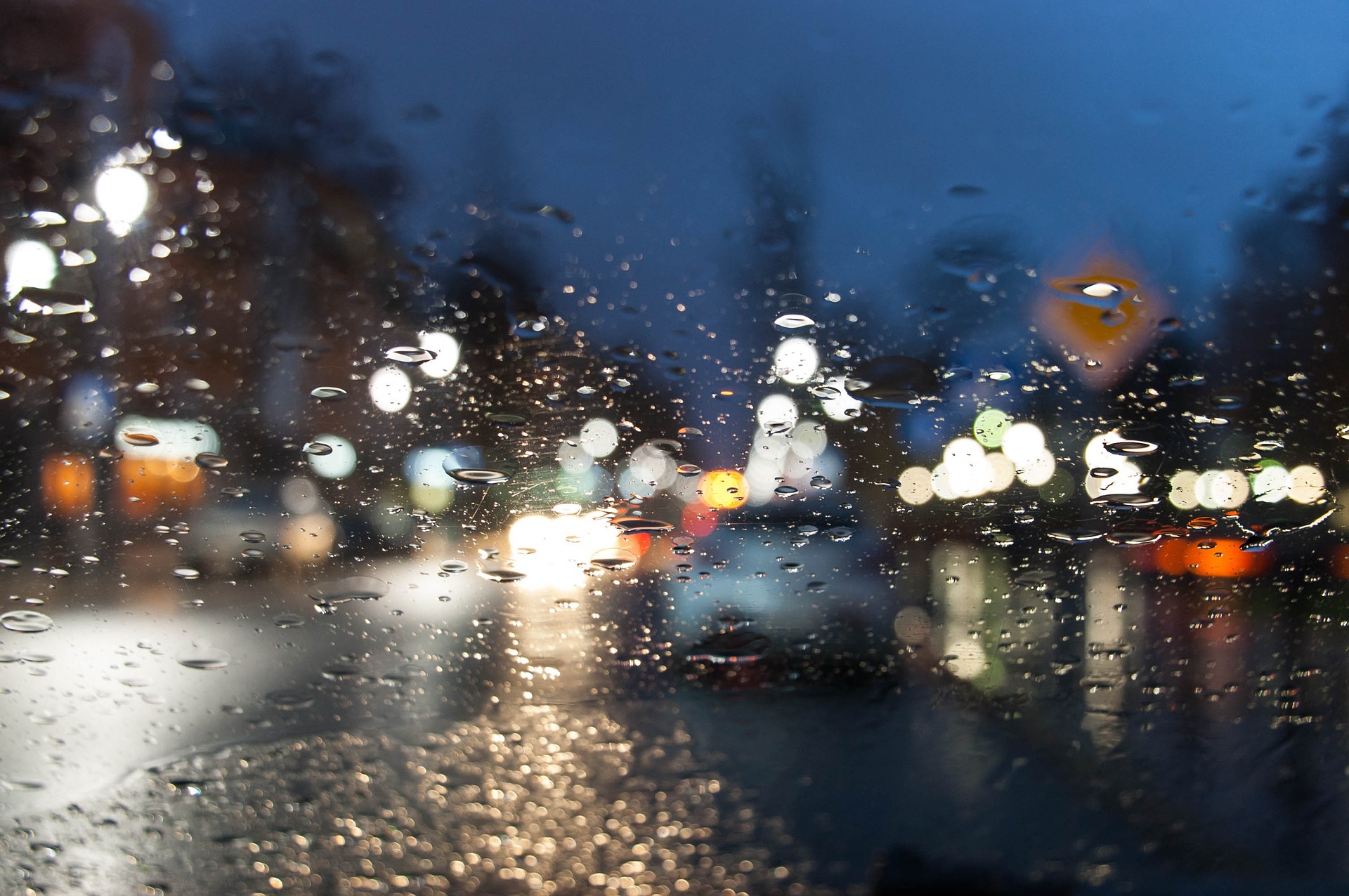 A nighttime scene viewed from inside a car, with raindrops scattered across the windshield. The raindrops create a blurred effect, distorting the colorful lights of the city outside.