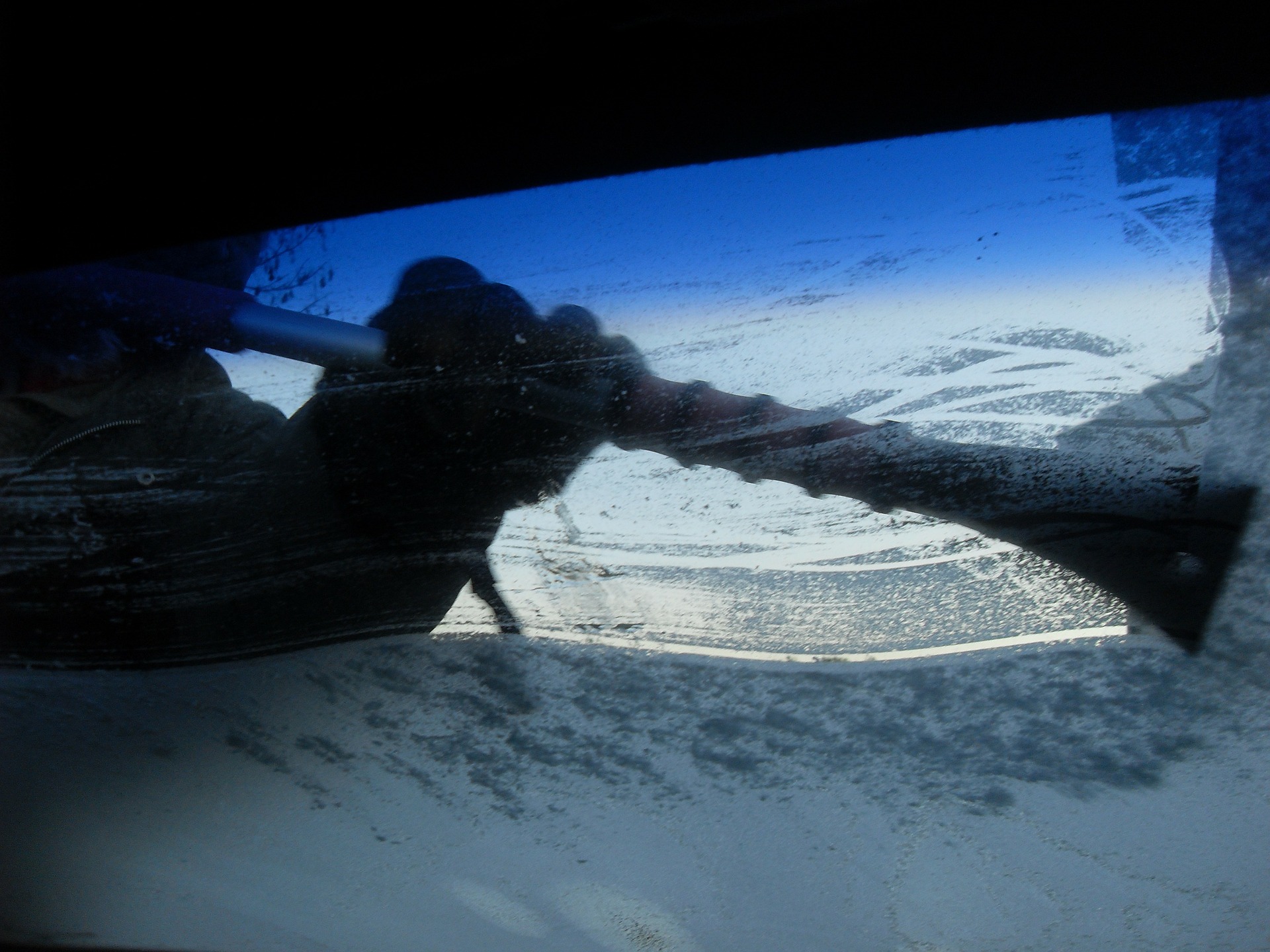 A person scraping ice off the windshield of a car from the outside, viewed from the interior of the vehicle. The image captures the gloved hand of the person holding a scraper and making sweeping motions to clear the frosty glass. 