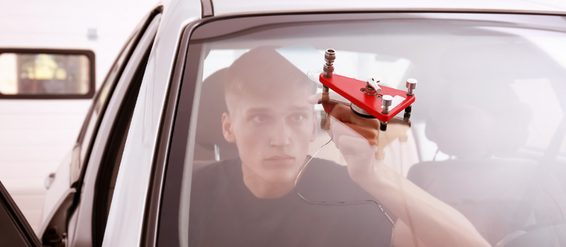 A technician is seen from inside a car, focusing intently on repairing a chip in the windshield. He is using a red windshield repair tool, which is attached to the glass with suction cups. The technician's concentrated expression highlights the precision and care required for the repair process. The interior of the car is partially visible, with the driver's seat and dashboard in the background. The scene takes place in a well-lit environment, possibly a garage or workshop, emphasizing the professional service being performed.