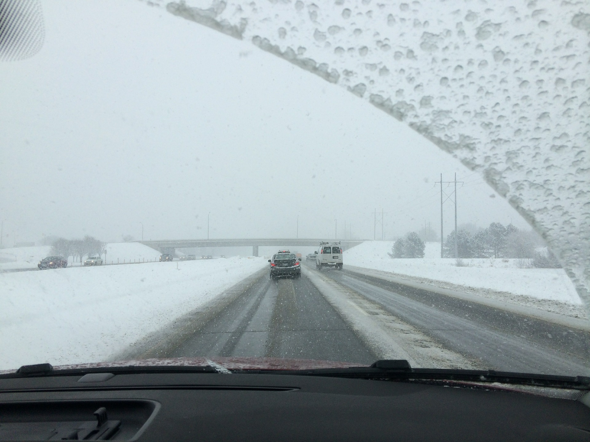 A view from inside a car driving on a snowy highway during a winter storm. The windshield is partially cleared, with the wiper blades leaving streaks of visibility through the snow and ice buildup. Several vehicles, including a van and a car, are ahead on the road, navigating the snow-covered lanes.