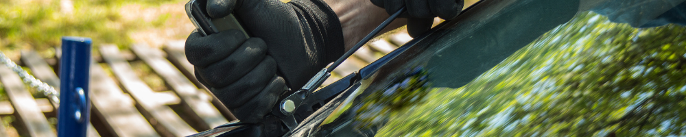 Close-up image of a person wearing black gloves and using a tool to cut through a car windshield, with blurred green trees reflected on the glass.