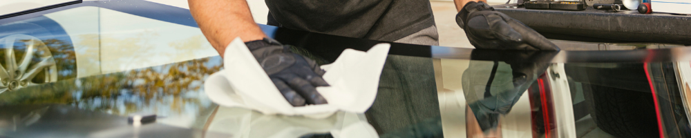 A person wearing black gloves cleaning the edge of a car windshield with a white cloth, with a car door open in the background and the reflection of trees visible on the glass.