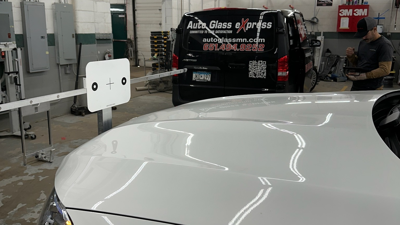 A white vehicle is positioned beside an ADAS calibration target inside an automotive glass workshop while a technician checks a handheld device. A black service van behind it displays the text "Auto Glass eXpress".