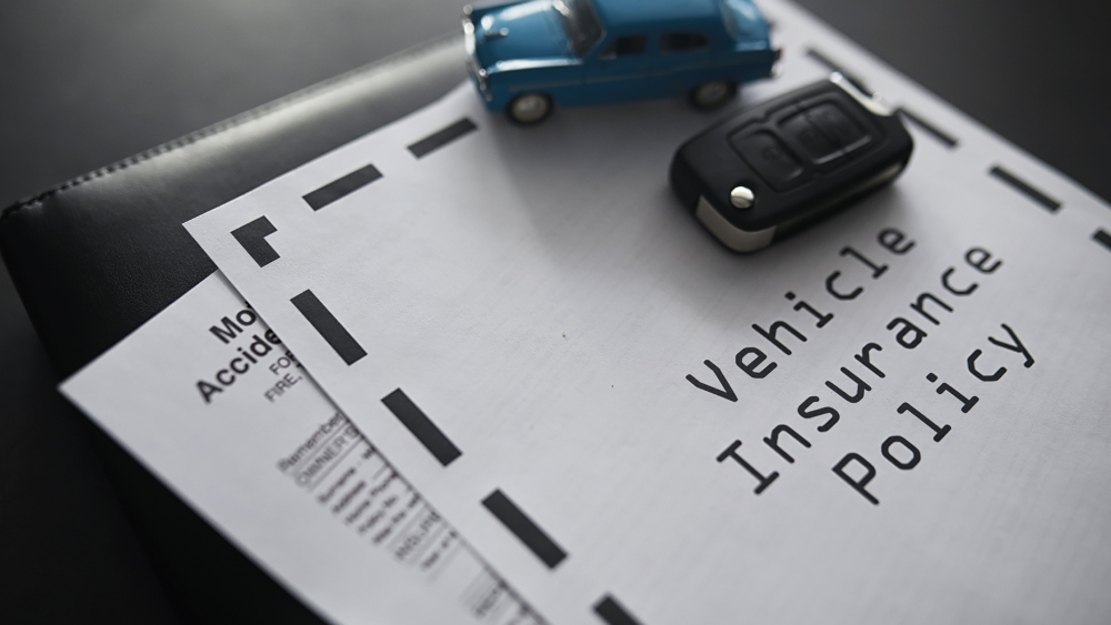 A close-up image of a vehicle insurance policy document placed on a dark surface. On top of the document are a small blue toy car and a black car key fob, symbolizing vehicle ownership and insurance coverage. The document's title, & Vehicle Insurance Policy is prominently displayed in bold black text. Partially visible beneath the insurance policy is another document related to motor accidents. The scene is well-lit, highlighting the importance of securing proper insurance for vehicle protection.