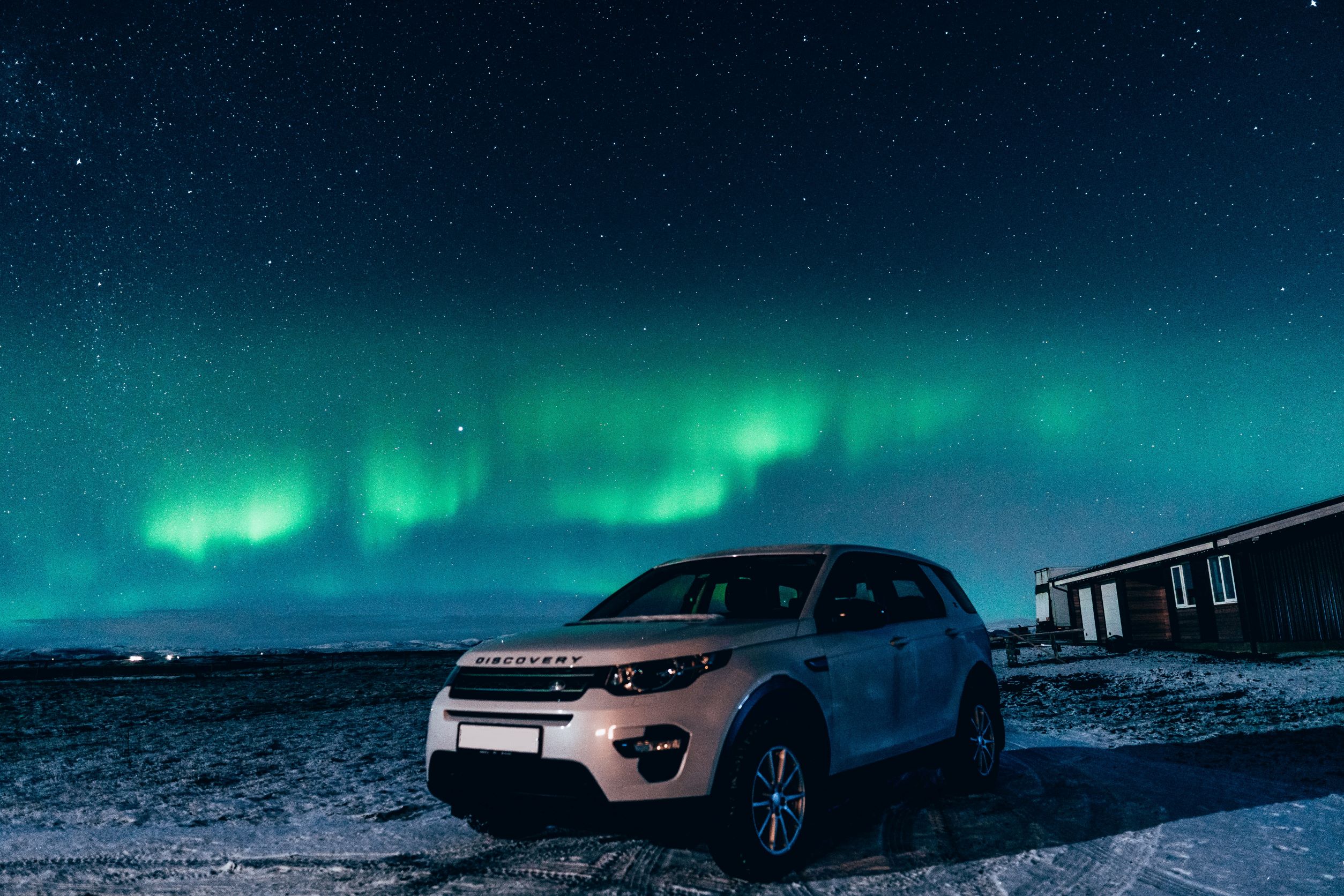 A Land Rover Discovery parked on a snowy terrain at night, with the aurora borealis visible in the starry sky above, next to a dark structure that resembles a house or shed.