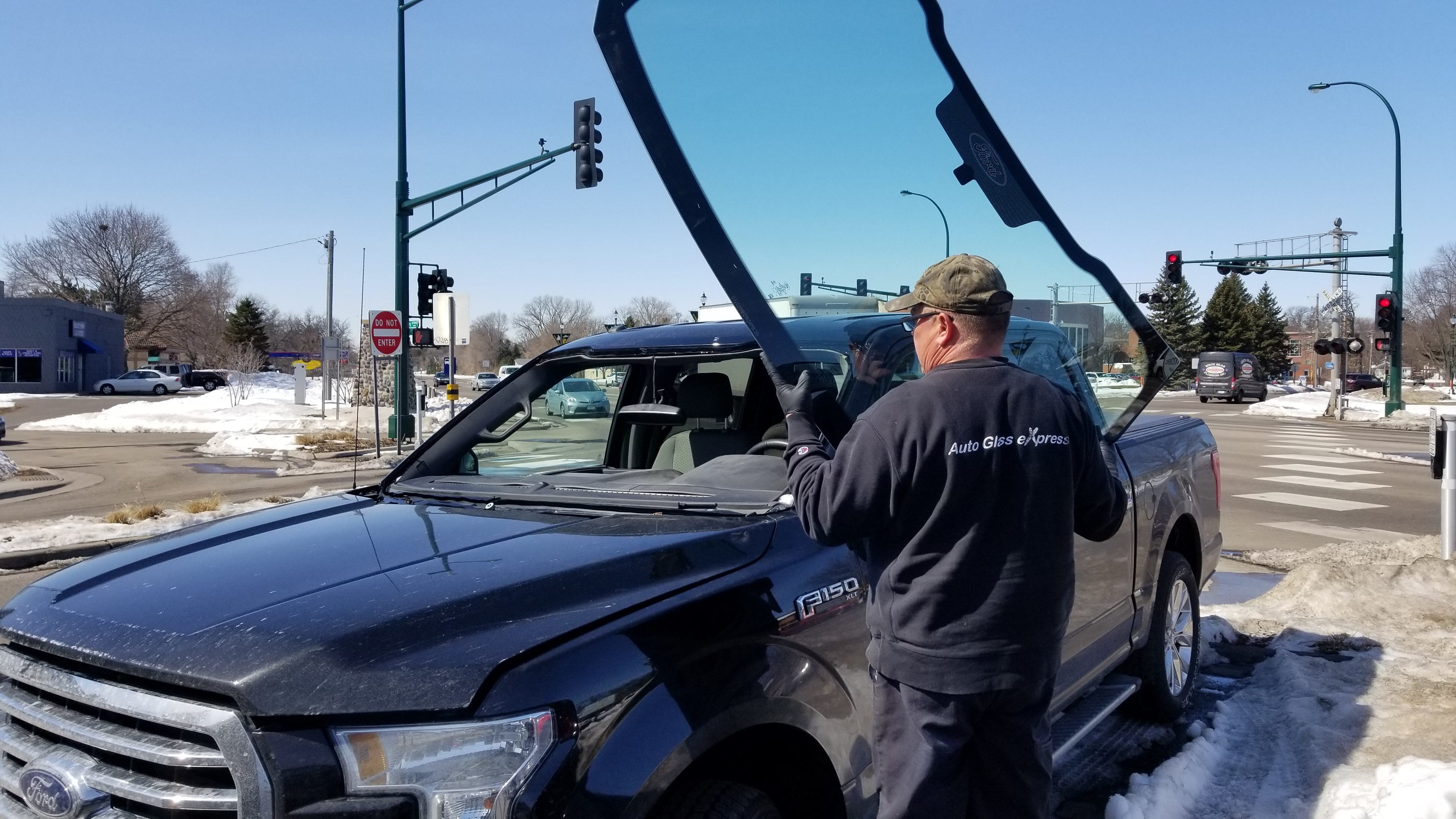 A technician from Auto Glass Express is installing a new windshield on a black Ford F-150 truck in White Bear Lake, Minnesota. The technician, wearing a cap and black work clothes, is carefully positioning the windshield into place.