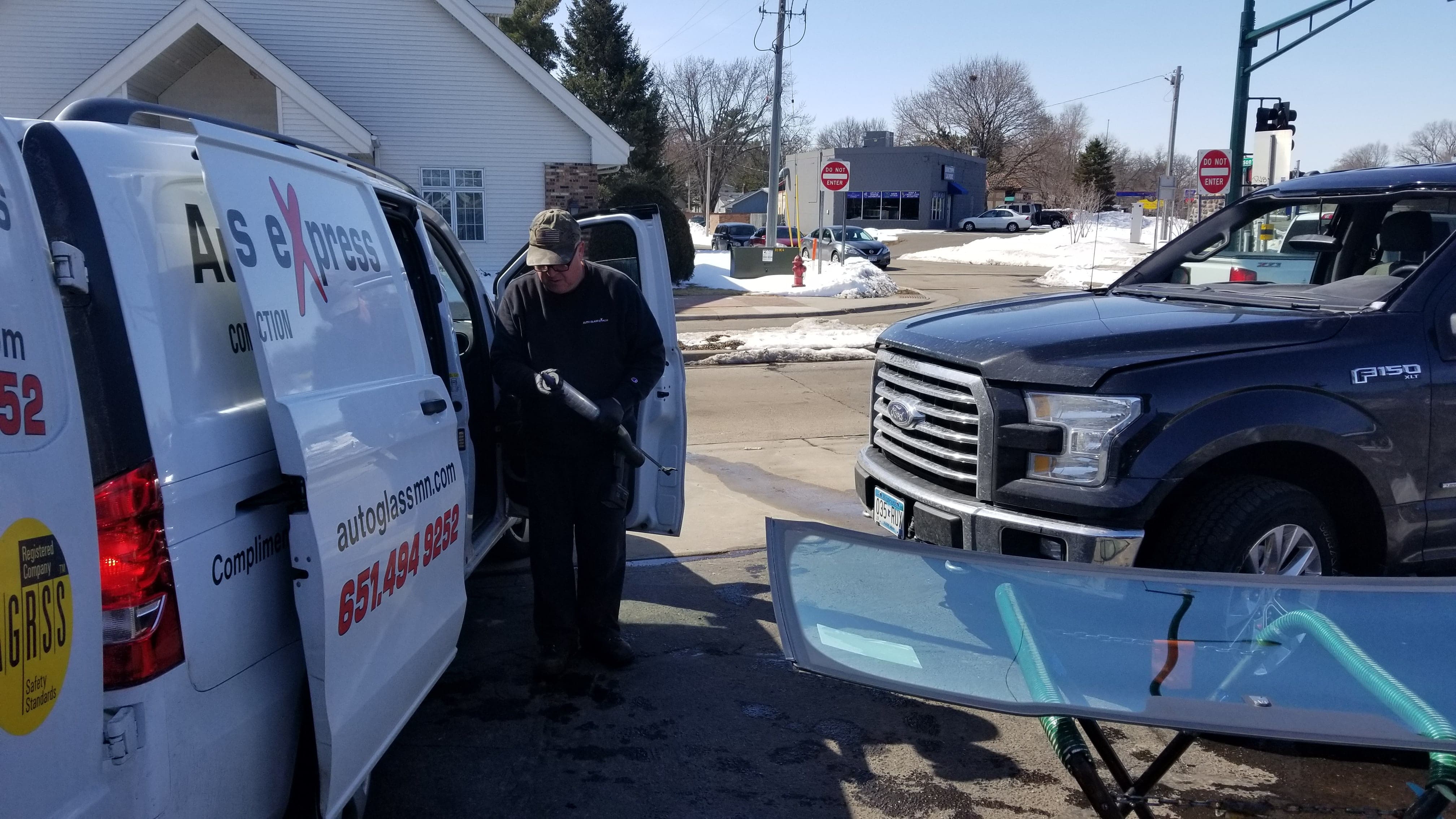 A technician working on a windshield replacement in White Bear Lake, MN. The technician, wearing a cap and black work clothes, is standing next to a white van with the logo 'Auto Glass Express' on the side. In the foreground, a new windshield is laid out, ready to be installed. The setting is an outdoor location with some snow on the ground and a mix of buildings and traffic signs in the background.