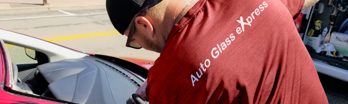 A man wearing a red shirt with the white text "Auto Glass eXpress" is working on a vehicle windshield repair in Blaine, Minnesota. The back of his shirt is visible as he leans over the windshield of the vehicle. He is wearing a black baseball cap and sunglasses, focusing intently on the repair of the damaged windshield. The image captures the professionalism and dedication of the auto glass service technician.