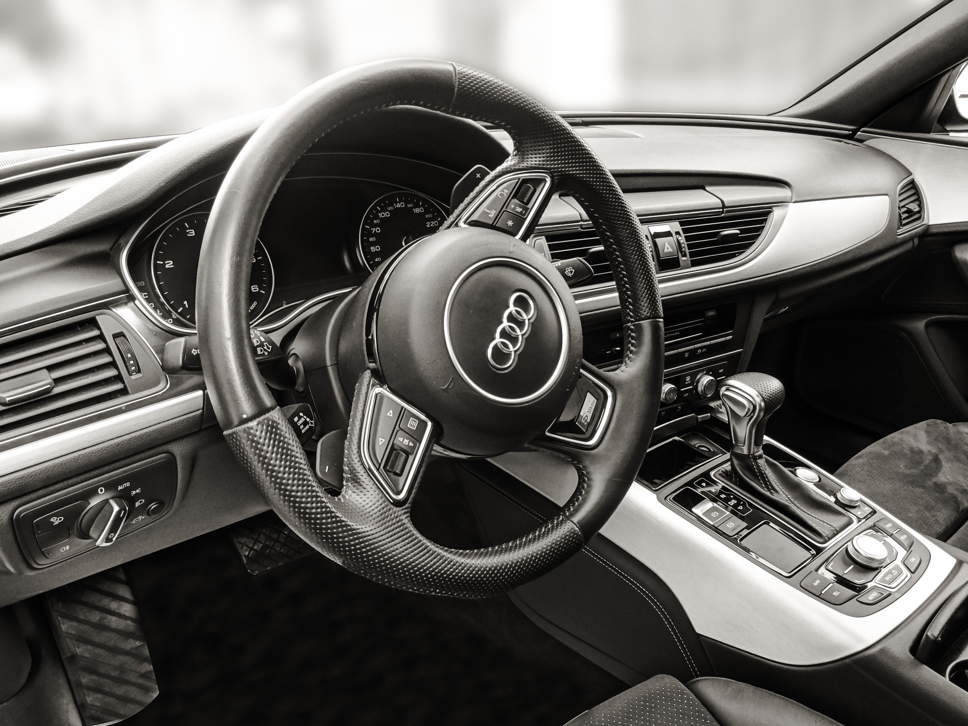 A detailed view of a modern car's interior, showcasing the driver's seat area. The black leather-wrapped steering wheel features the Audi logo at the center and various control buttons. Behind the wheel are two large, circular instrument gauges with clear, easy-to-read markings. The dashboard has a sleek, black and silver design, with air vents, an infotainment system, and climate control knobs prominently displayed. 