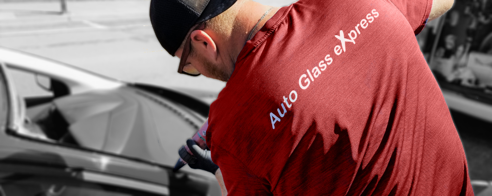 A man wearing a red shirt with the white text "Auto Glass eXpress" is working on a vehicle windshield. The back of his shirt is visible as he leans over the windshield of the vehicle. He is wearing a black baseball cap and sunglasses, focusing intently on the repair of the damaged windshield. The image captures the professionalism and dedication of the auto glass service technician.