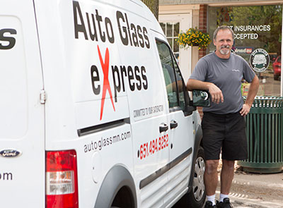 A man wearing a red shirt with the white text "Auto Glass eXpress" is working on a vehicle windshield repair in [city, state]. The back of his shirt is visible as he leans over the windshield of the vehicle. He is wearing a black baseball cap and sunglasses, focusing intently on the repair of the damaged windshield. The image captures the professionalism and dedication of the auto glass service technician.