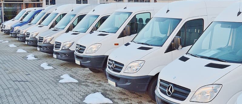 A lineup of white Mercedes-Benz vans parked in a row on a paved surface in the Twin Cities metro area. There are multiple identical vans positioned side by side, facing the same direction. Small patches of snow are visible on the ground in front of the vehicles, suggesting a cold or winter setting.